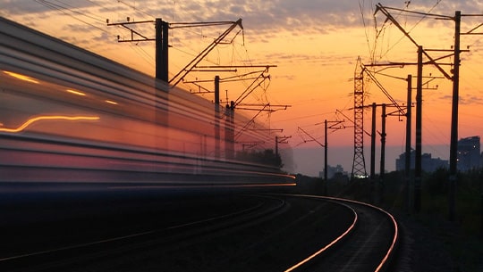 Das Bild zeigt Oberleitungen und einen fahrenden Zug vor einem Sonnenuntergang.