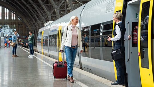 Das Bild zeigt eine Frau mit Koffer am Bahngleis vor einem Zug.