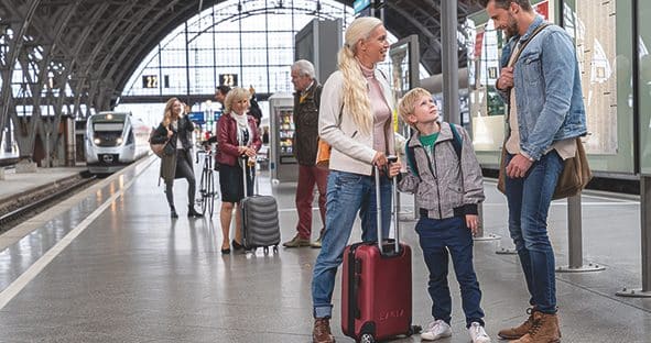 Das Bild zeigt Personen mit Gepäck-Rollkoffern an einem Bahnsteig.
